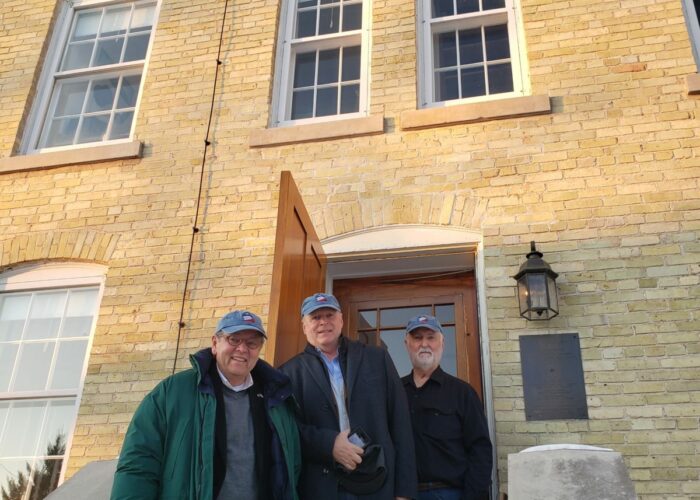 Three men standing in front of a brick building with date reading 1860.